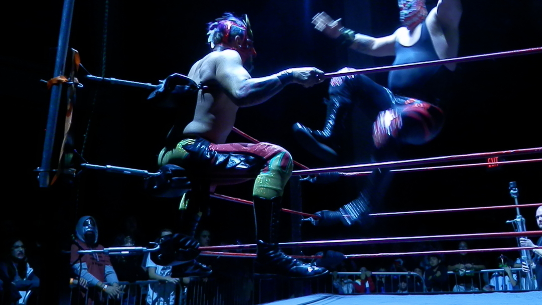 A photograph of two professional wrestlers in motion, one about to knee the other while he sits on the turnbuckle.